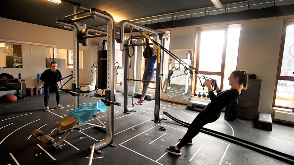 Drei Personen trainieren in einem Fitnessstudio, lächeln, schwarz gekleidet, mit Fitnessgeräten.