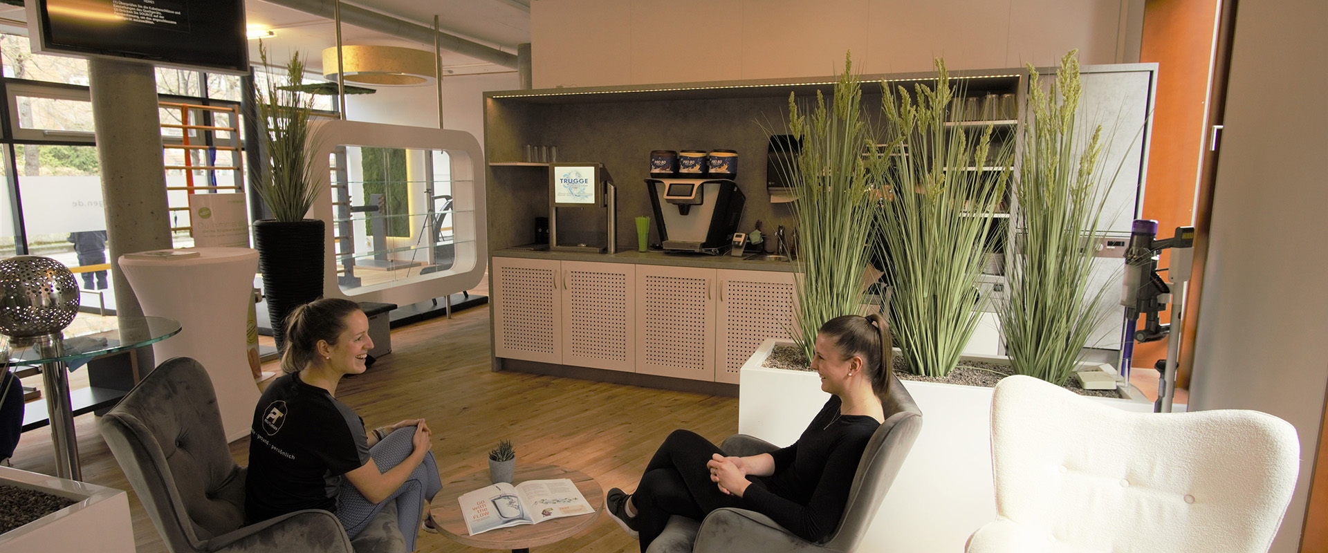 Zwei Frauen in moderner Lounge, lächelnd, helle Holzböden, Pflanzen, gemütliche Atmosphäre.
